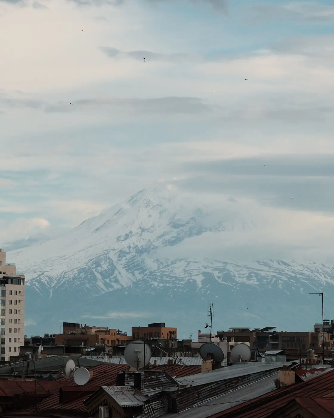 Yerevan rooftops with satellite dishes and tin roofs, Ararat under cloud behind