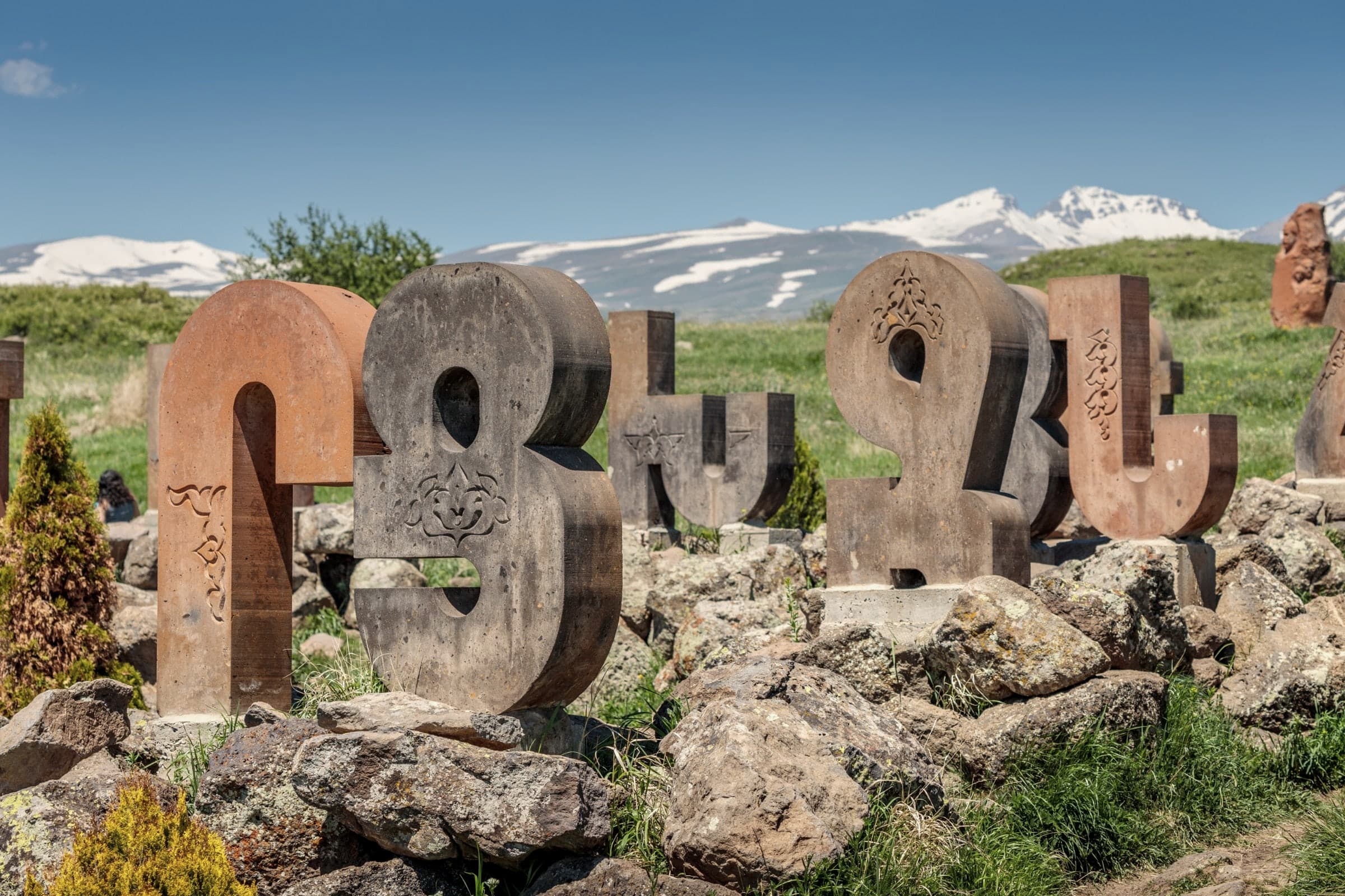 Carved tuff letters of the Armenian alphabet among rocks, the snow-streaked summits of Aragats behind