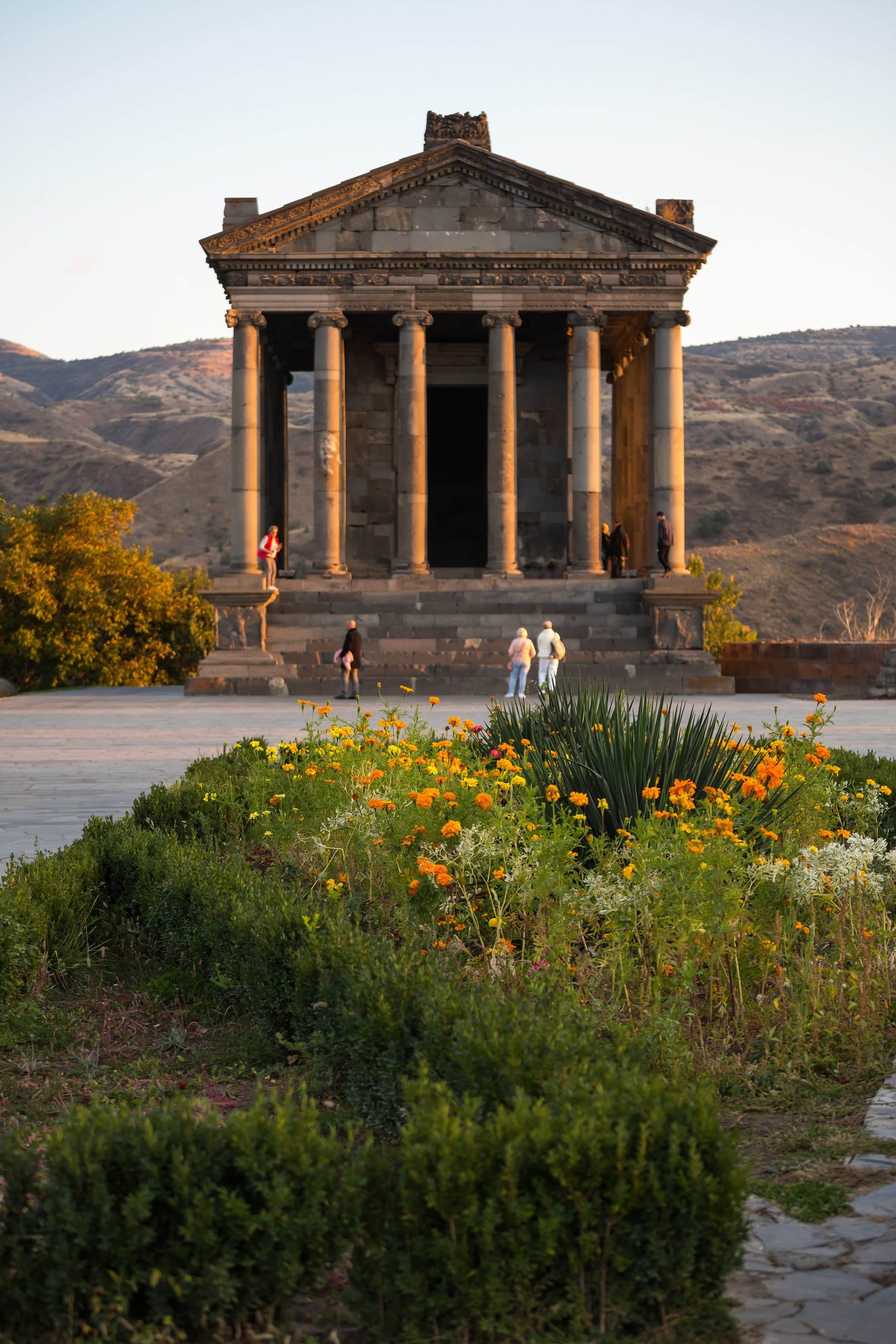 Garni temple from the front in warm low sun, a bed of marigolds in the foreground and brown hills behind
