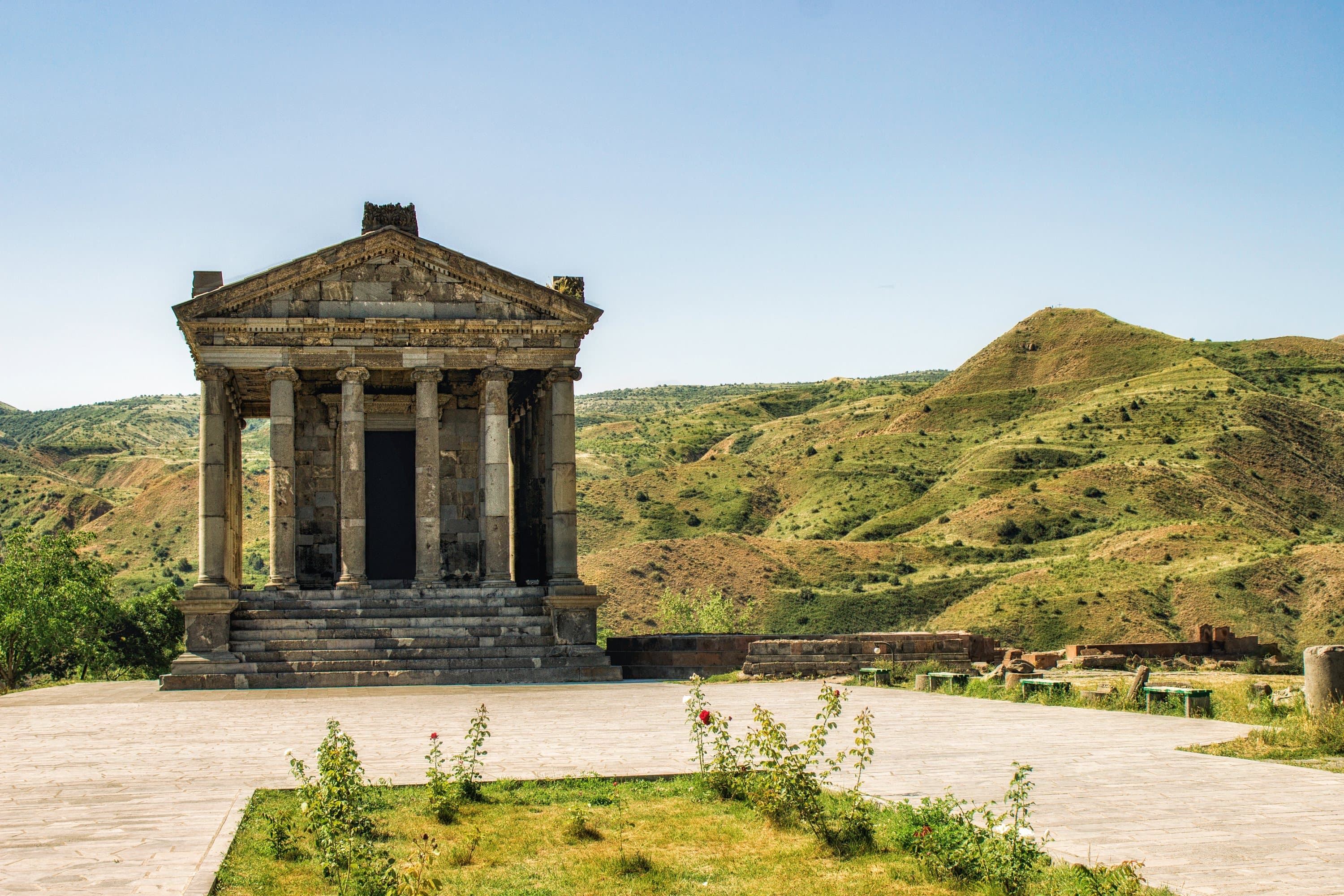 Garni temple in clear daylight on its spur, green and ochre hills rising behind under a pale violet sky