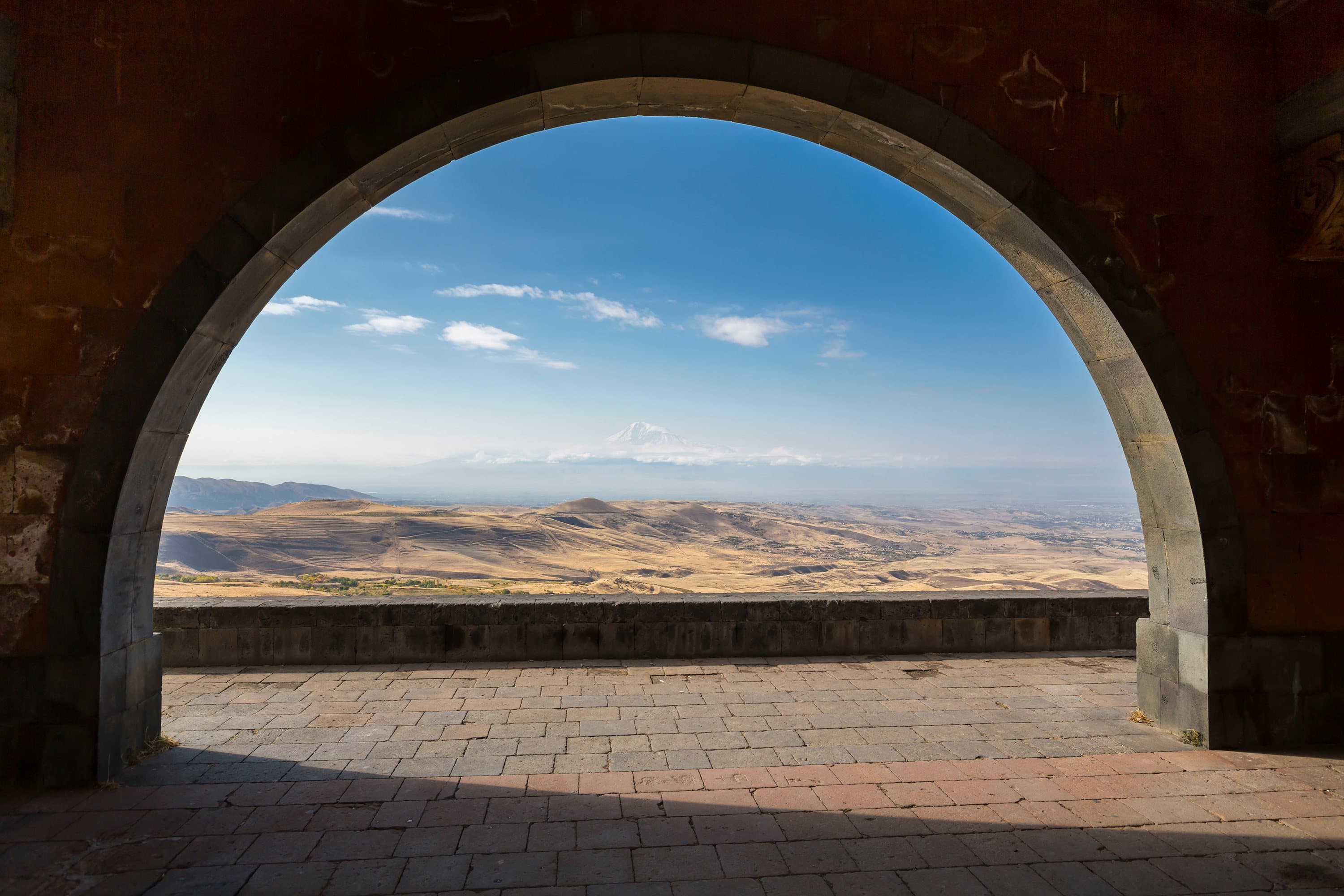 Ararat’s snow peak framed through the stone Arch of Charents, above the dry golden plain and a paved terrace