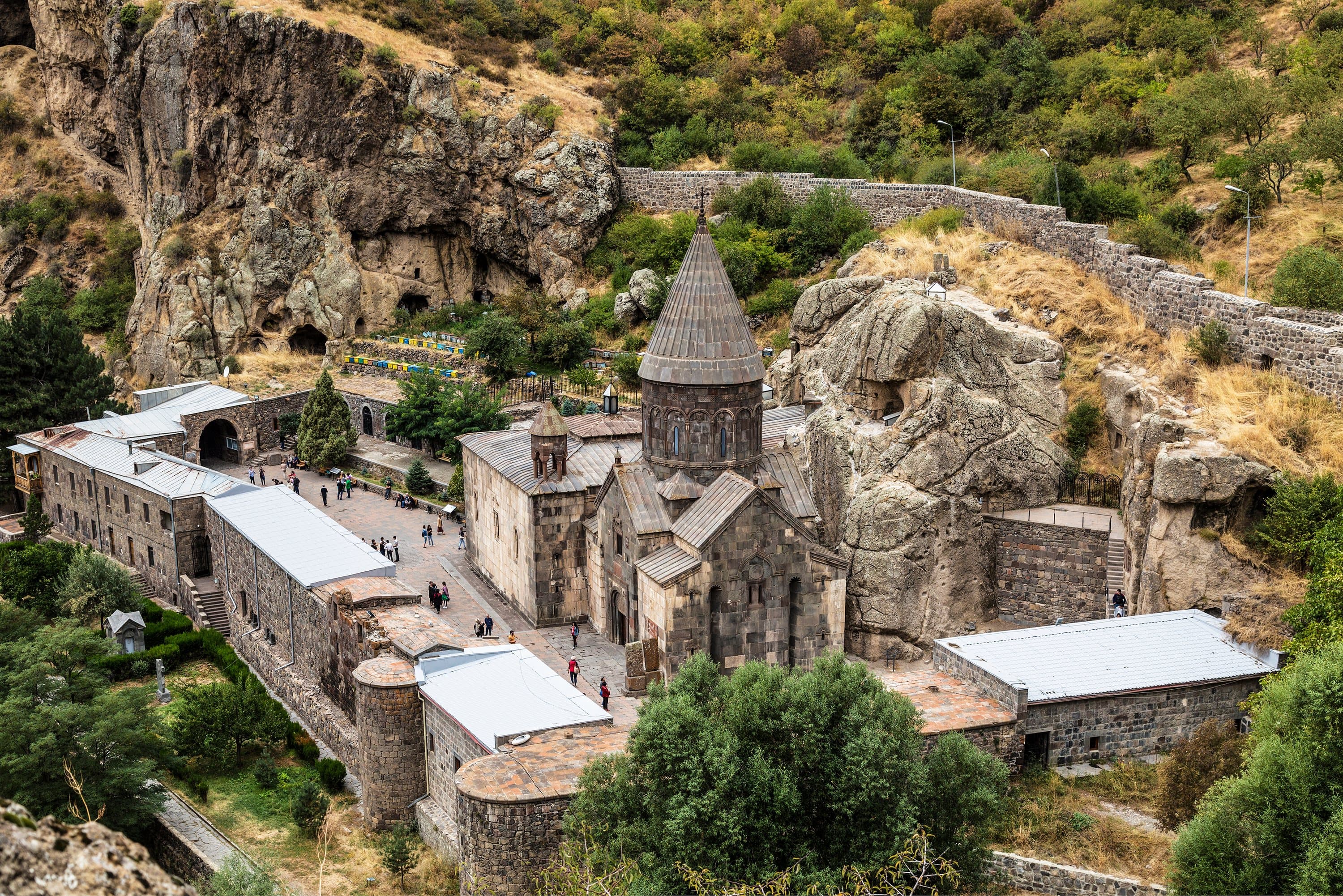 Geghard from above: the church and walled courtyard set hard against the cliff, with rock-cut openings in the rock face behind.