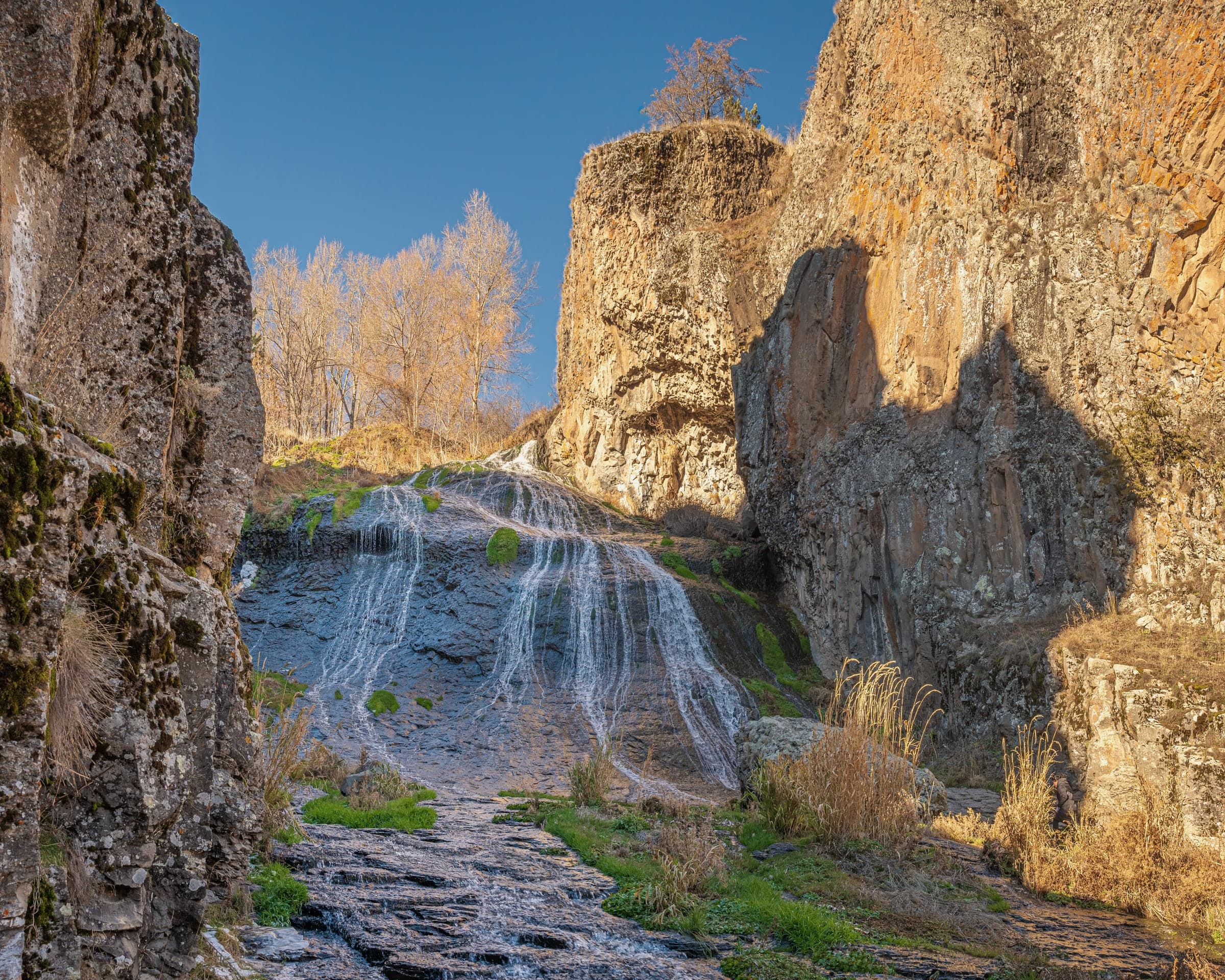 The Jermuk waterfall spreading in thin streams over dark rock between basalt walls, bare trees gold on the rim in autumn light