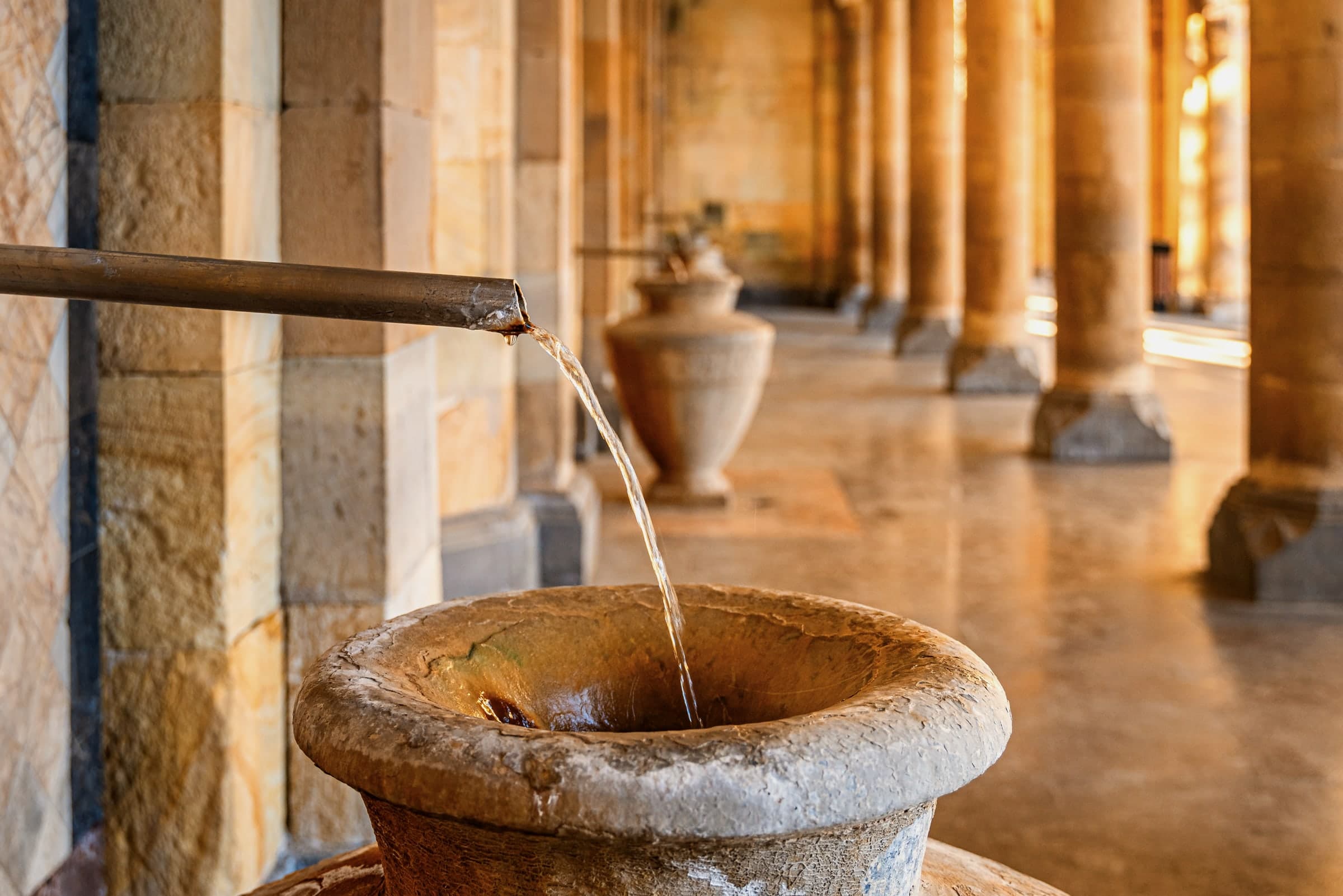 Mineral water pouring from a pipe into a stone urn in Jermuk's gallery of waters, a colonnade of sandstone piers behind