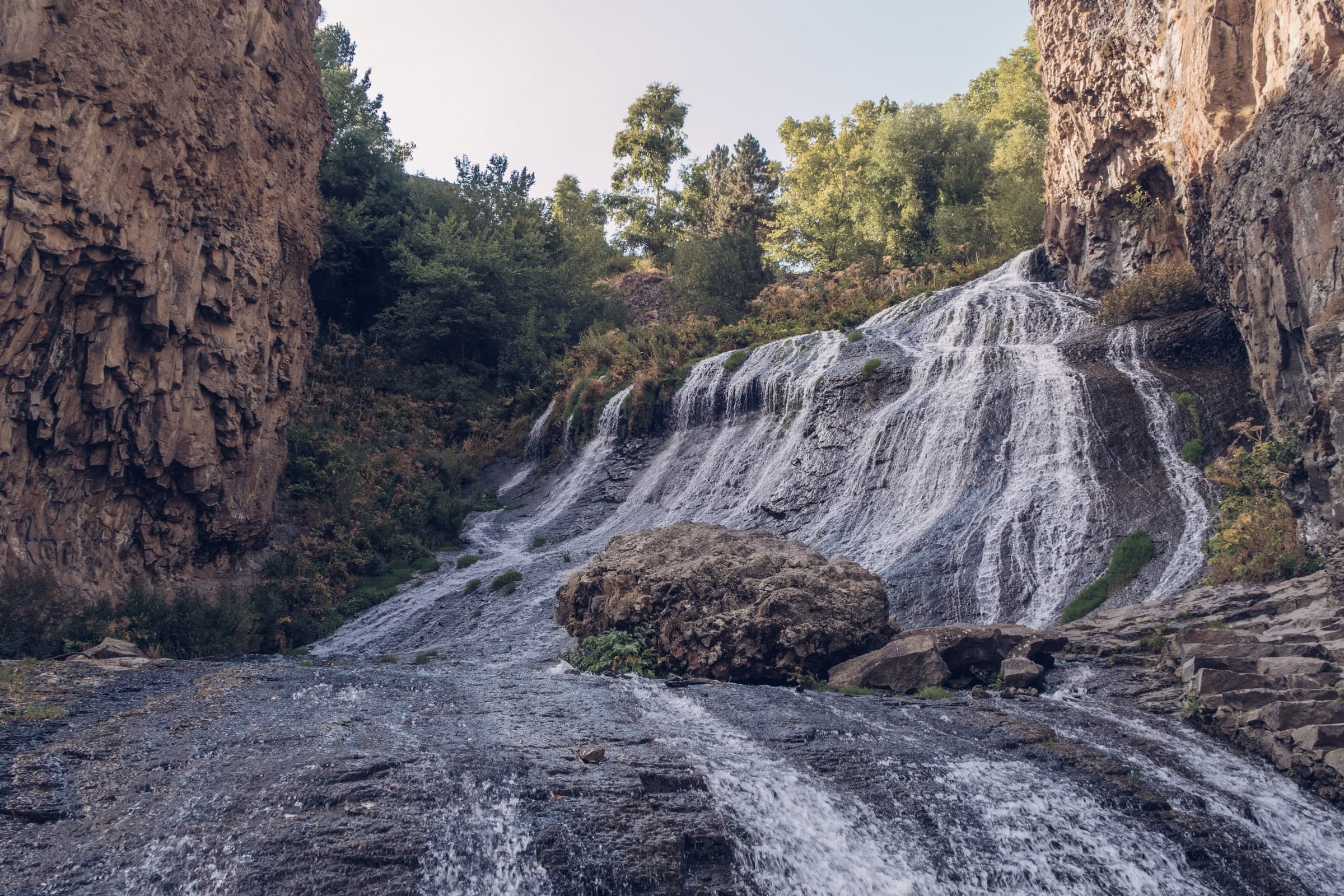 The foot of the Jermuk waterfall in summer, water fanning over the rock past a boulder, trees and cliffs above