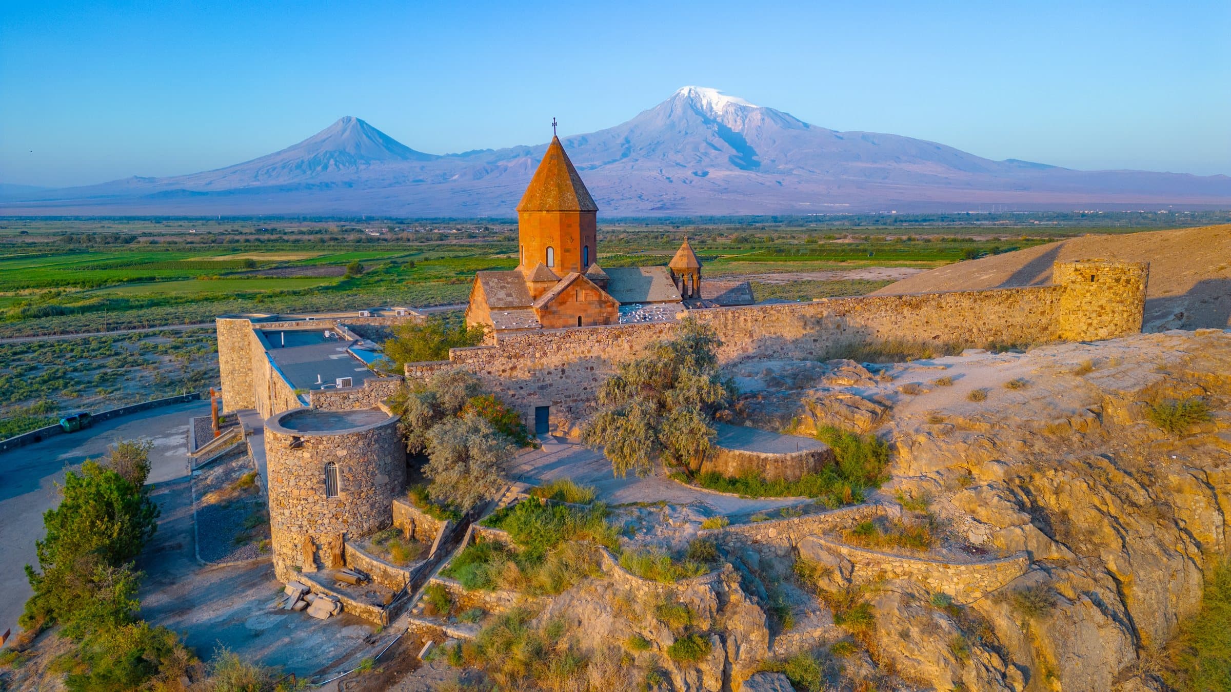 Khor Virap from above in early light, its church and walls on the rock, with both peaks of Ararat across the plain