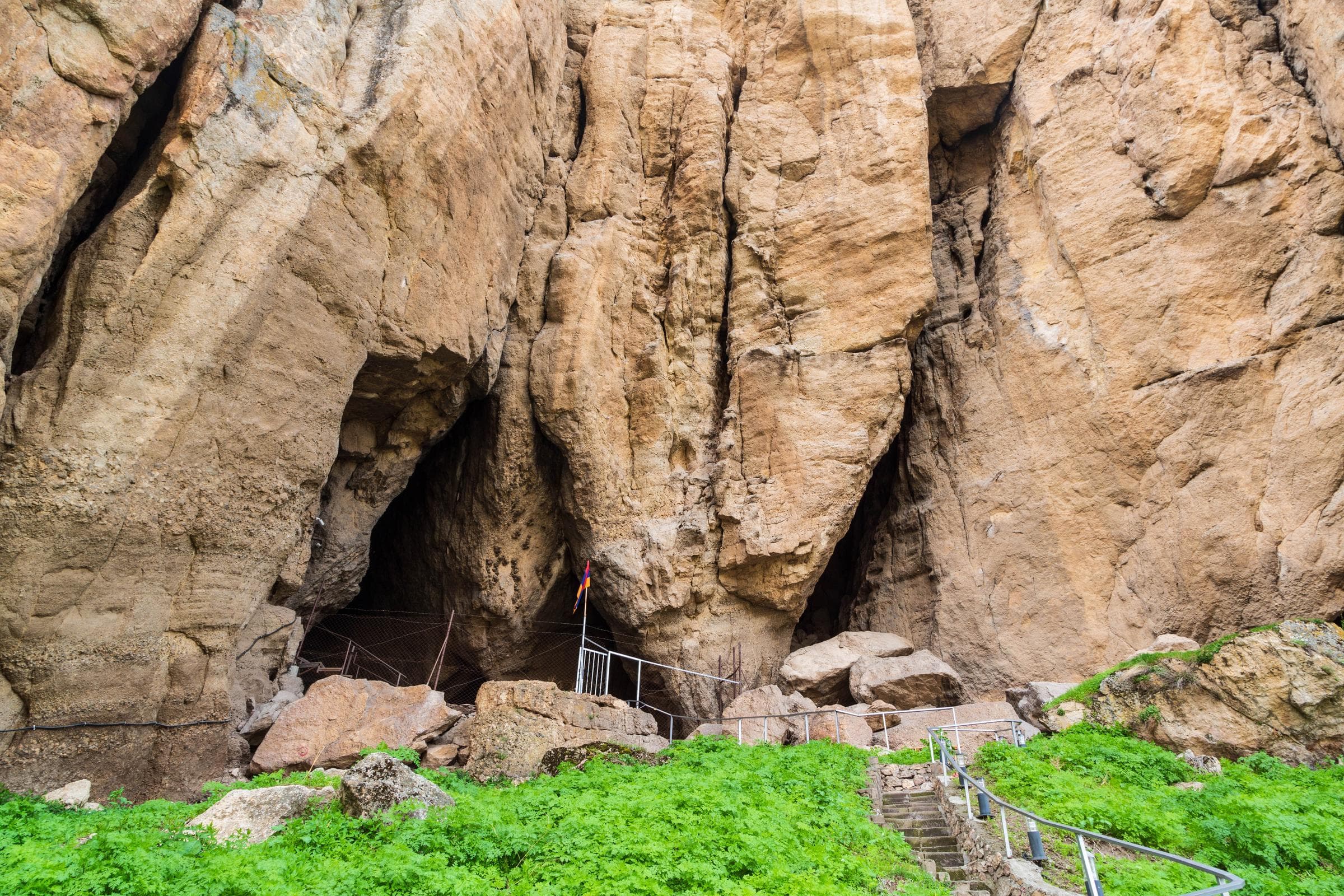 The mouths of the Areni-1 cave in a wall of pale rock, reached by a railed stair through green ground