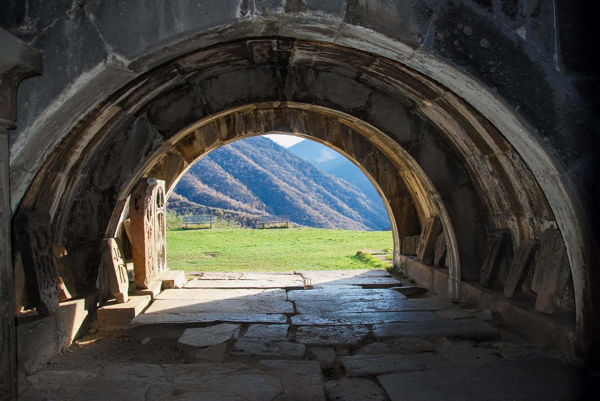 A vaulted stone passage at Haghpat with carved cross-stones, opening onto a green slope and the mountains