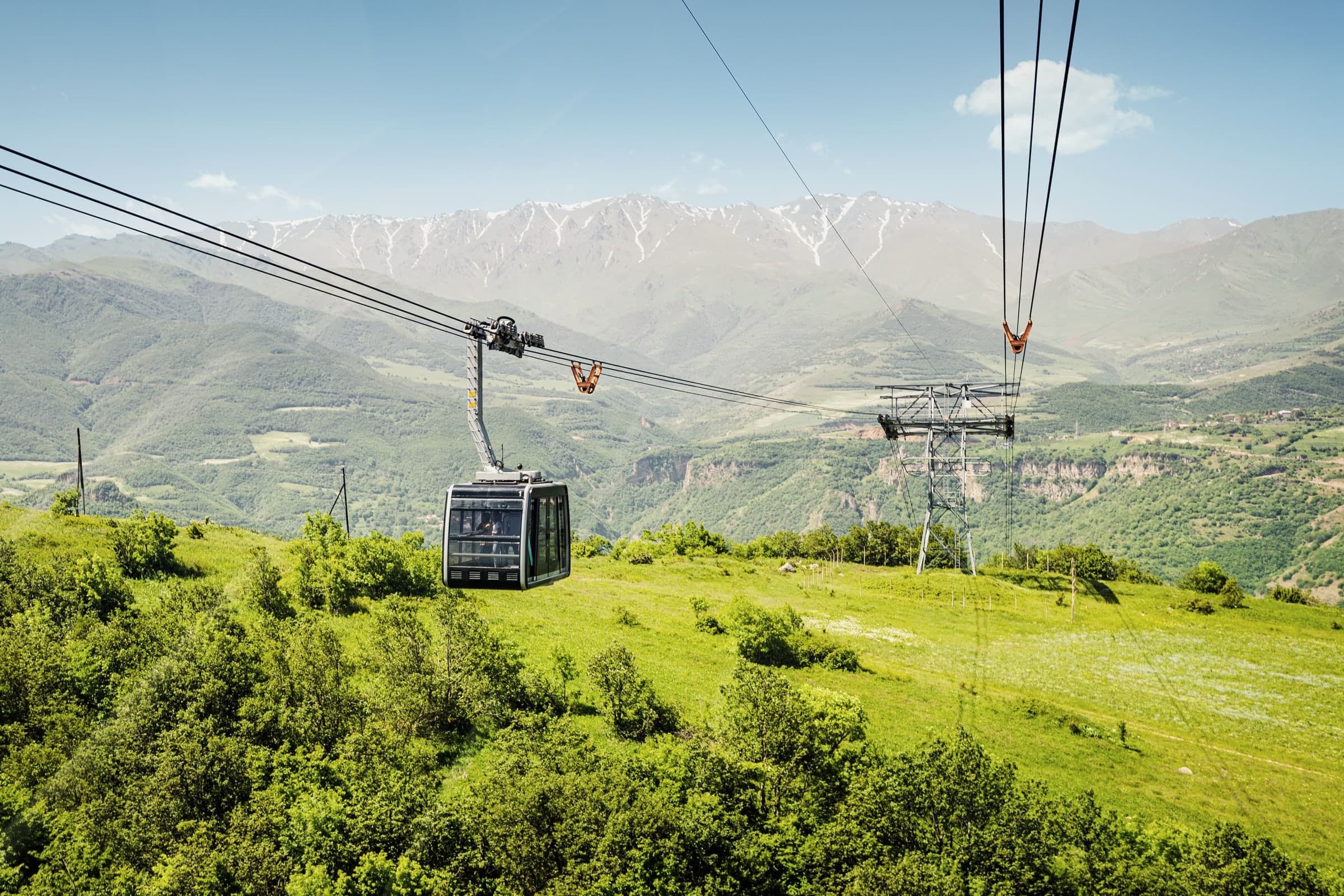 A Wings of Tatev cabin above green slopes, snow-streaked mountains behind