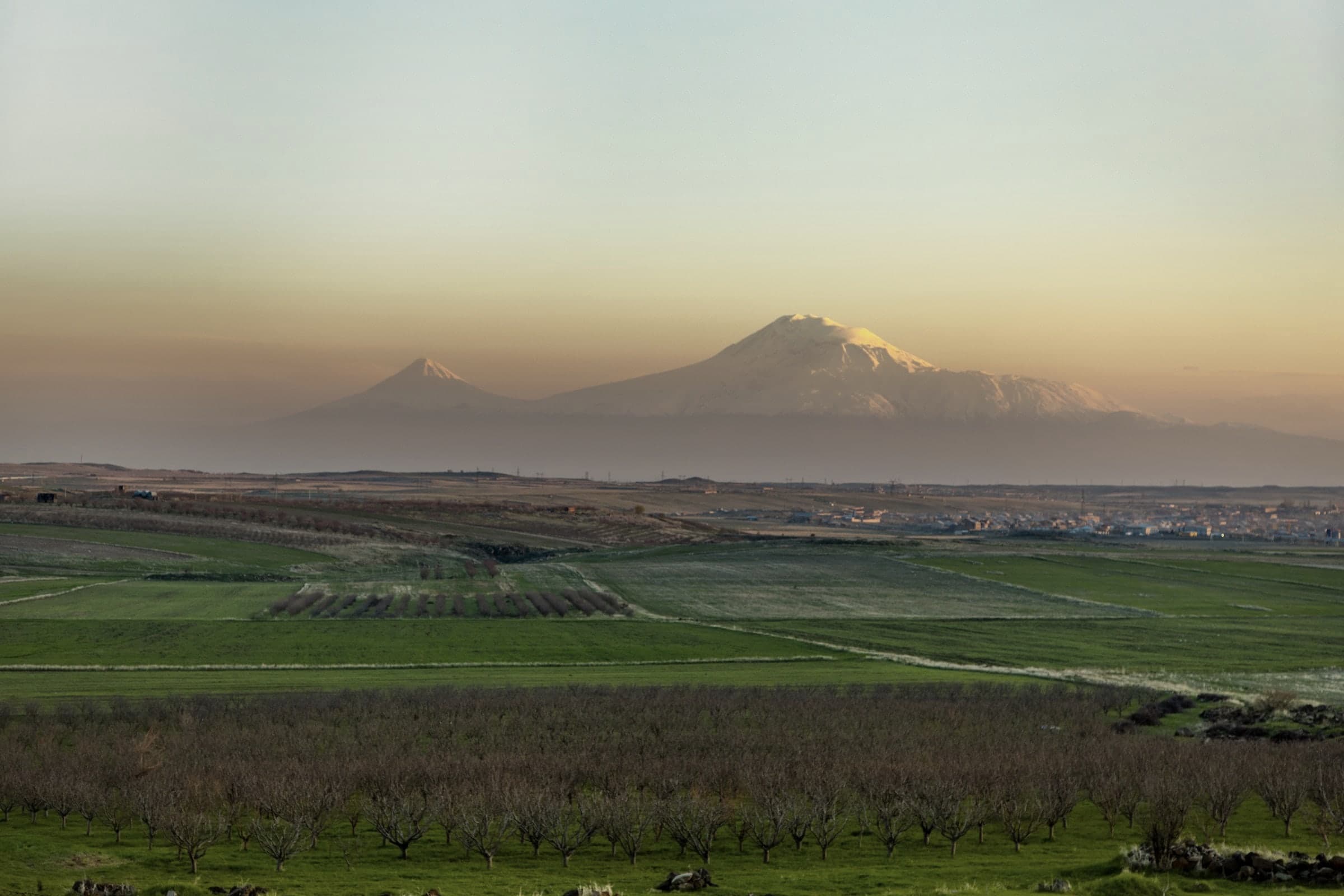 The Ararat plain at sunset: green fields and orchard rows, a village on the right, and both peaks of Ararat rising out of the haze