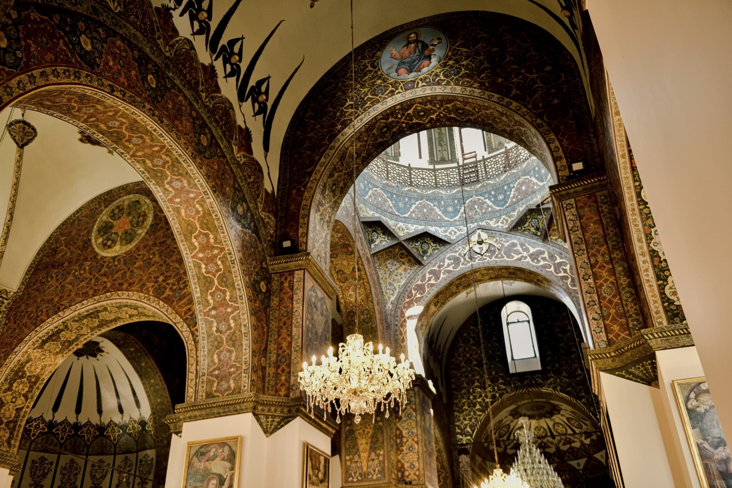 Inside Echmiadzin cathedral: painted arches in red and gold, a blue painted dome above, a lit chandelier below