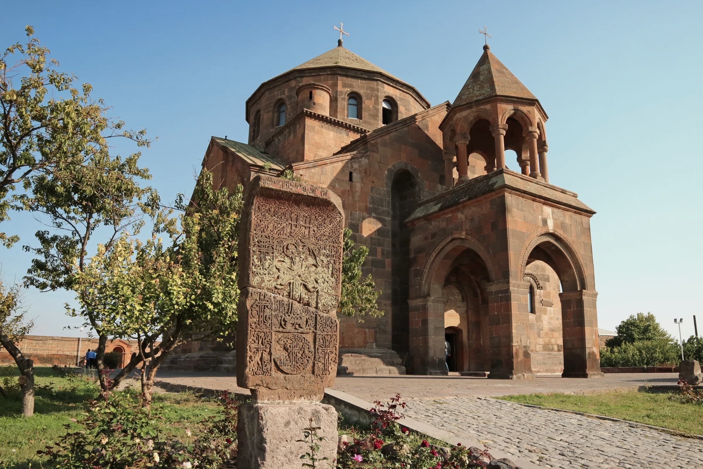 St Hripsime in warm late light, a carved khachkar in front and a cobbled path leading to the door