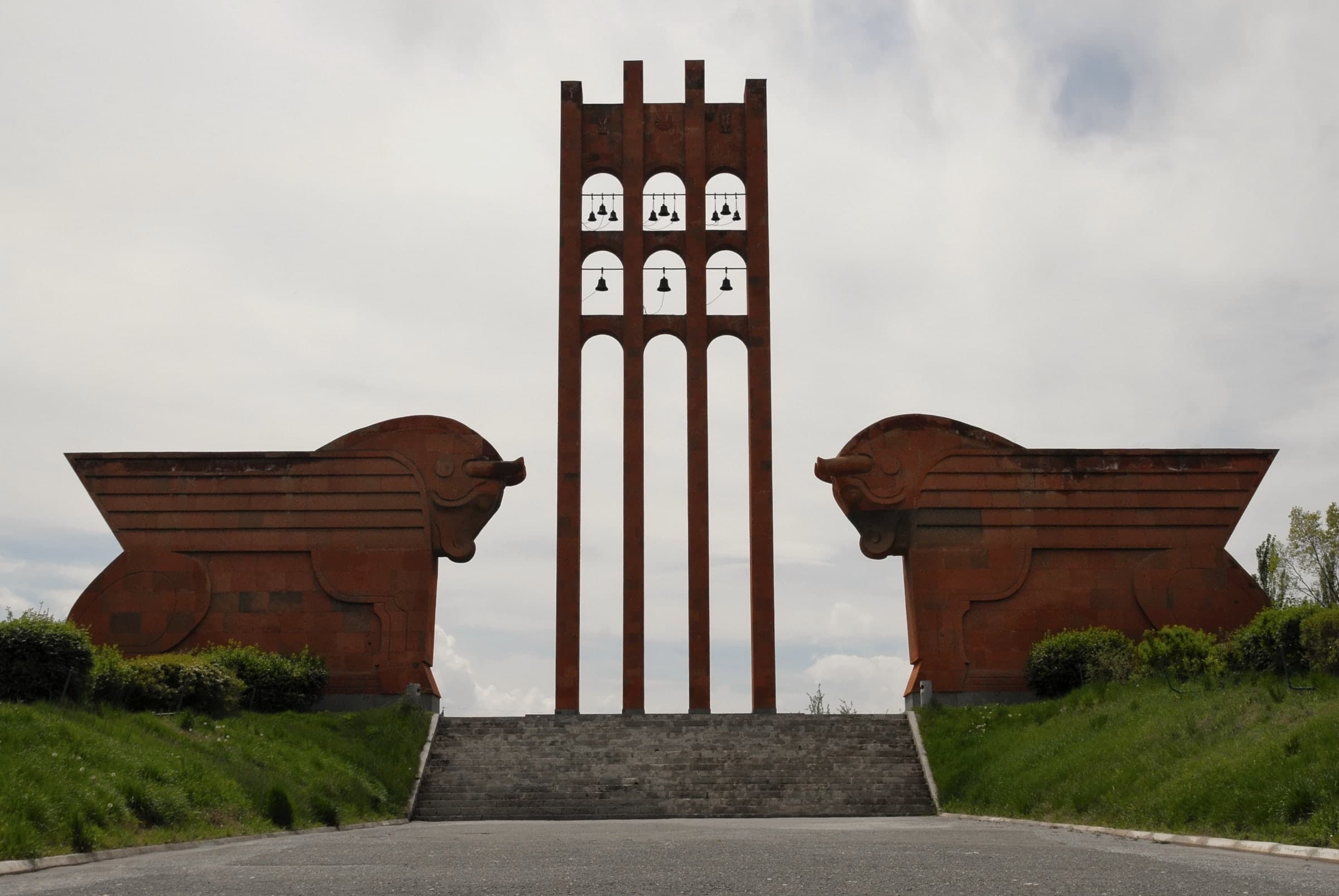 The Sardarapat memorial: a tall red-tuff bell tower between two giant winged bulls at the top of a flight of steps, under an overcast sky