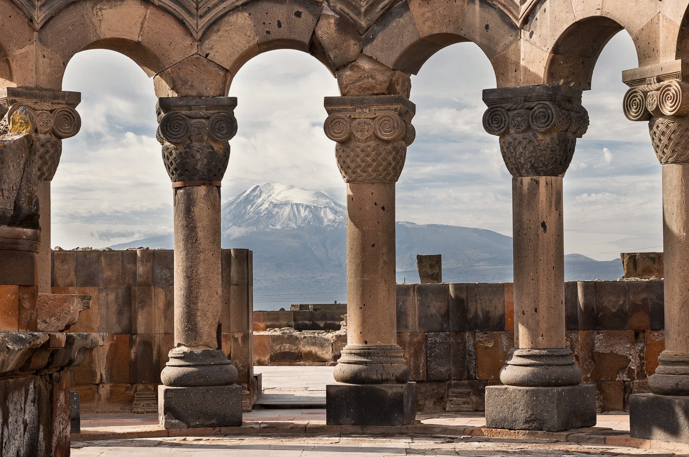 Inside the ring at Zvartnots: carved capitals with spirals and woven basketwork on tuff columns, Ararat framed in the arch between them