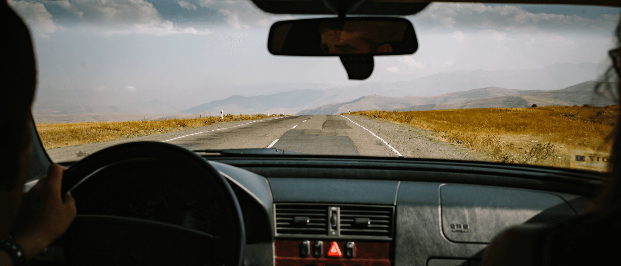 The view through a windscreen: an empty two-lane road across dry golden steppe, mountains hazy ahead, hands on the wheel