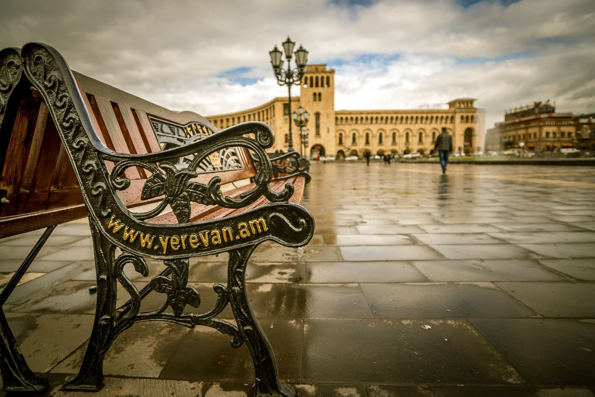 A wet Republic Square after rain, a cast-iron bench in the foreground and the arcaded tuff facades behind